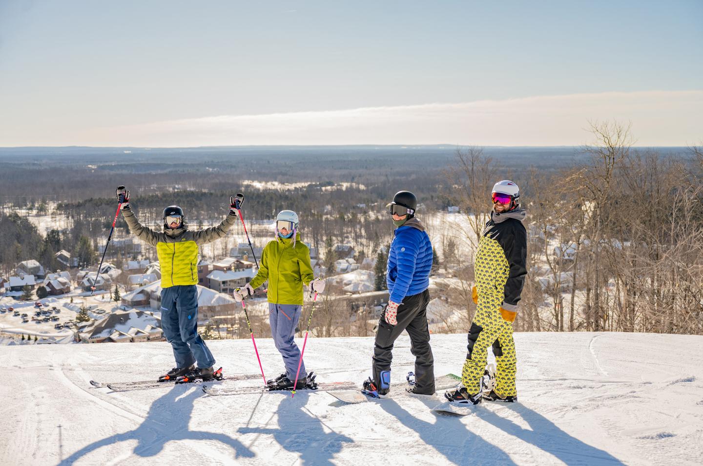 Four skiers in colorful outfits atop a snowy hill, with distant wintry landscape.