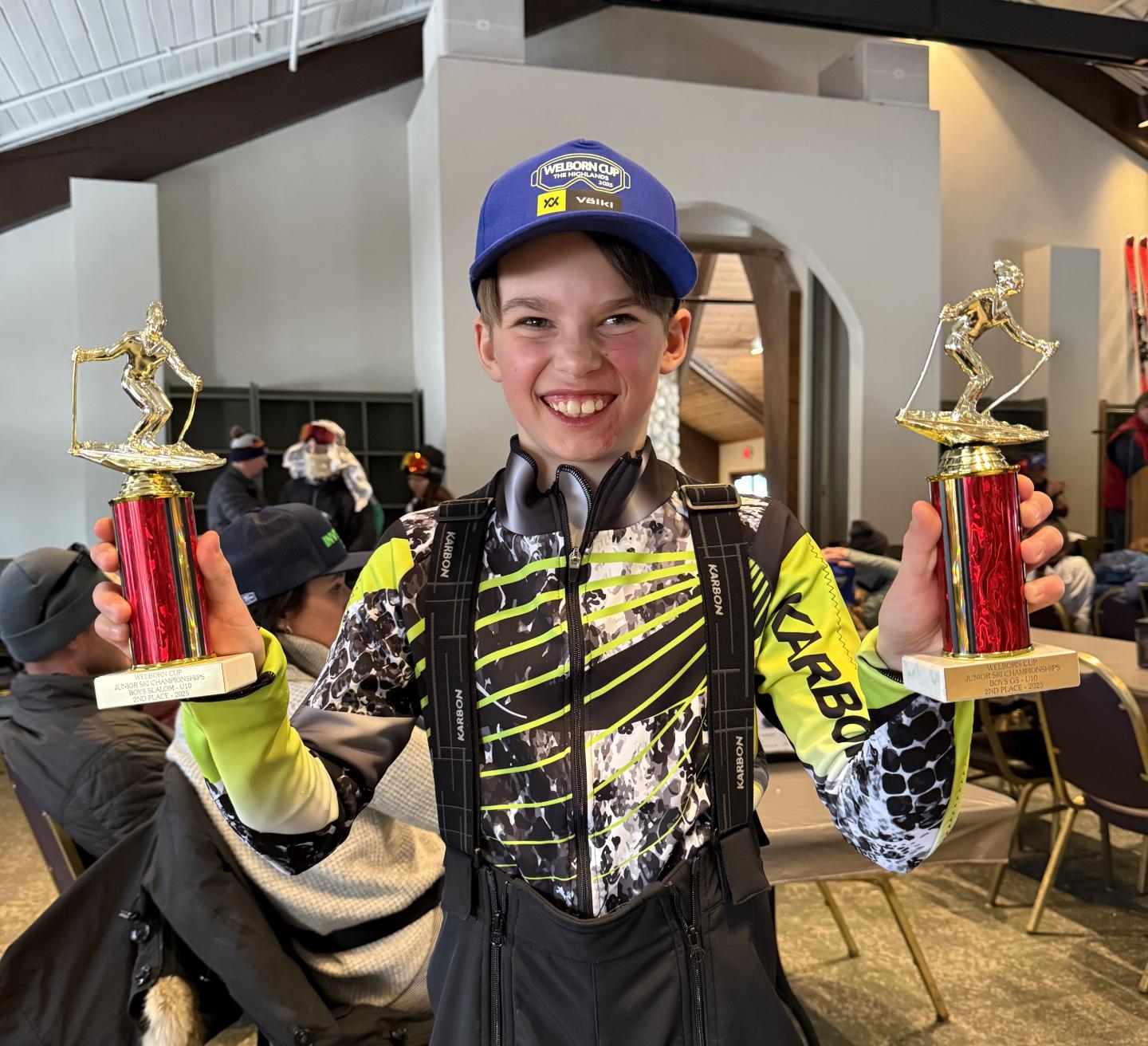 Smiling child holding two trophies indoors, wearing a blue cap.