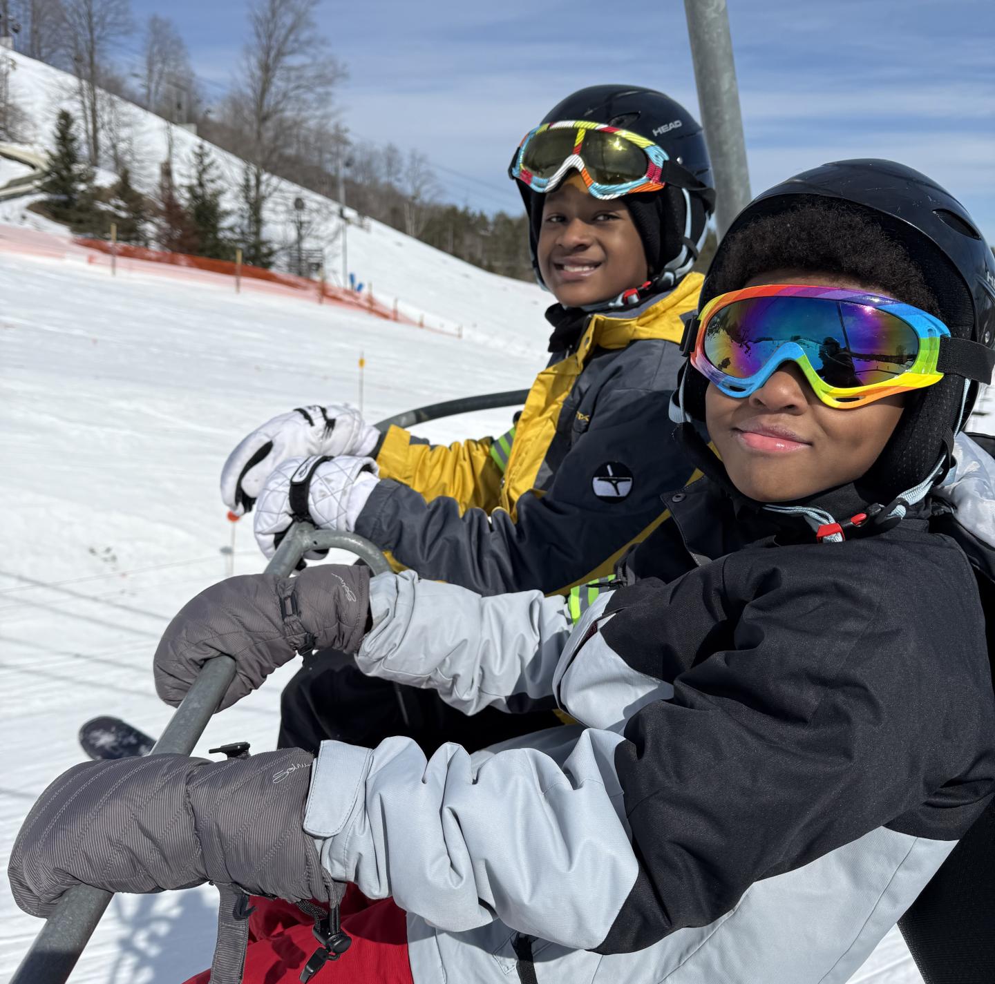 Two skiers in helmets and goggles on a ski lift, snowy landscape in the background.