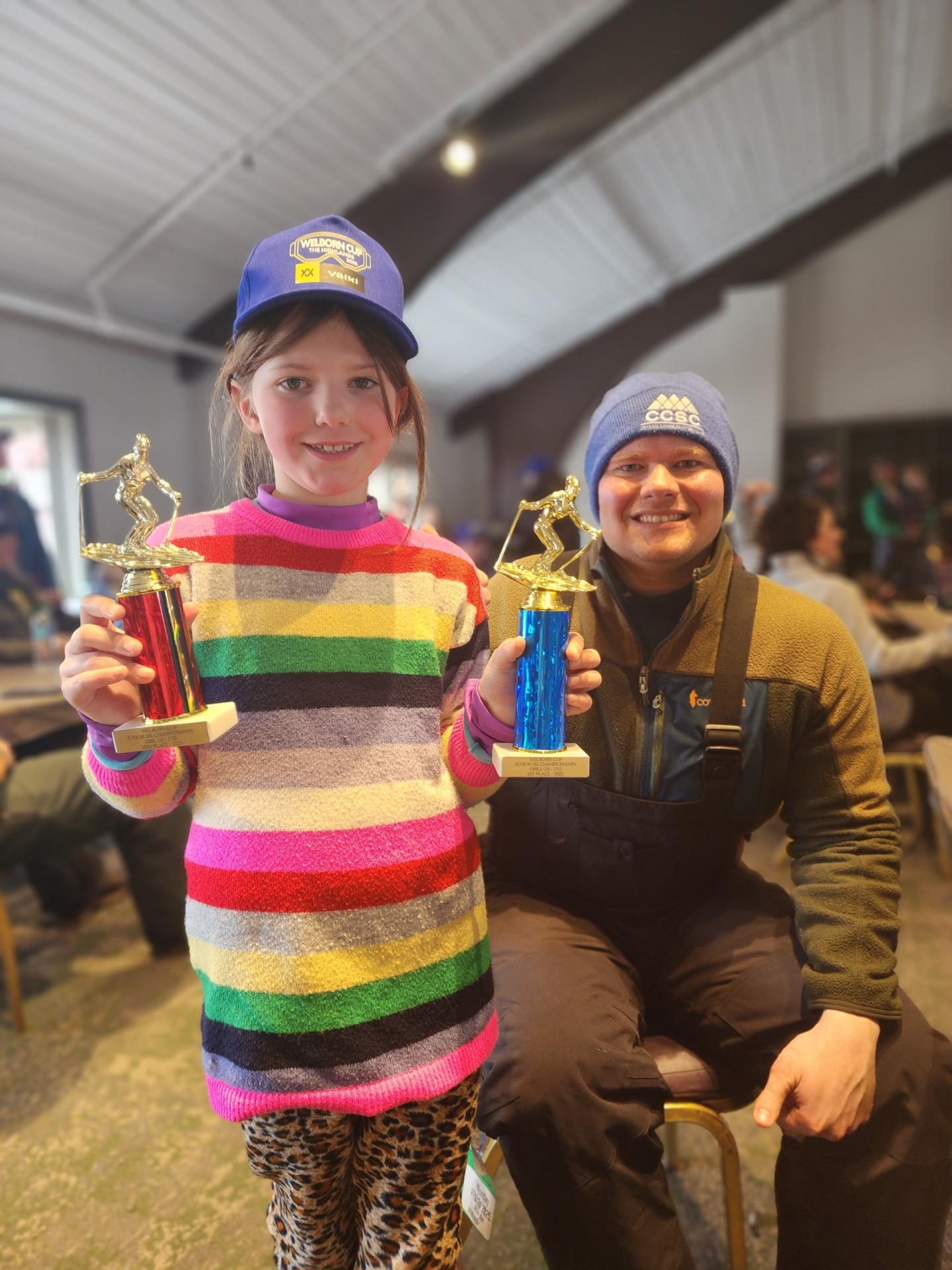 Child and adult smiling, holding trophies indoors. Colorful striped outfit, both wearing hats.